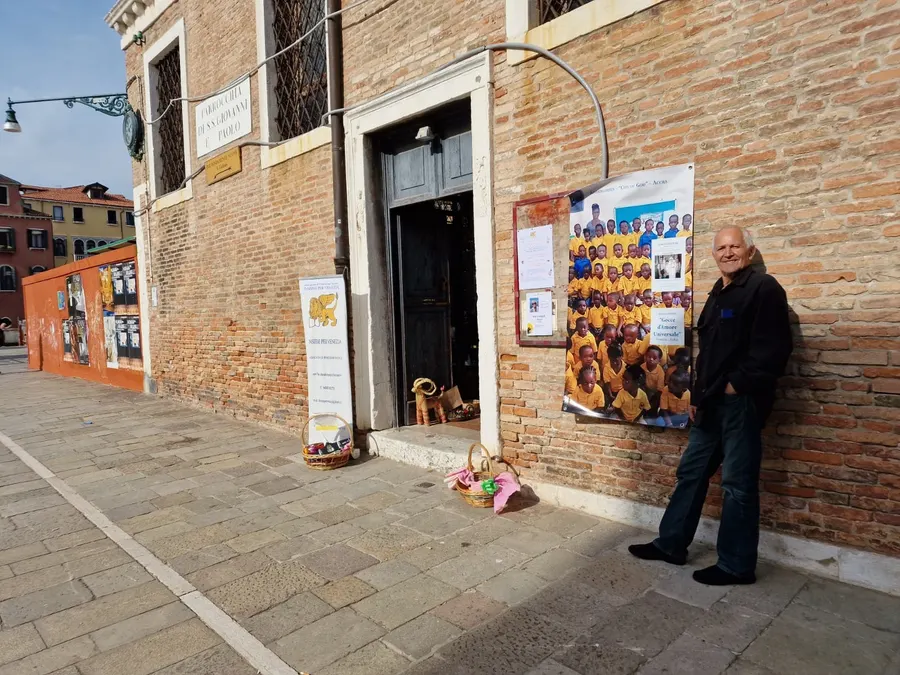 Claudio accanto al banchetto del mercatino solidale davanti a una facciata in mattoni a Venezia